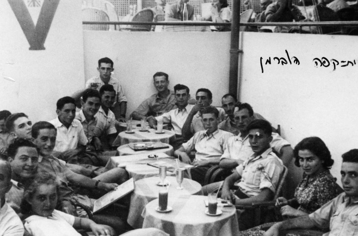 פלוגה ד', תיאור התמונה: חופשה בתל-אביב, קפה הוברמן. מופיעים בתמונה: מימין: משה רונן, יפה גולדברג, ברהרב, ישראל שטריקס, שמואל רוזנפלד, מומה, ד.אריאלי, עמיאל, משה גולדפרב, אהרון ידידיה, ישעיה ברוט, ברוך יקותיאלי, רפאל רקנטי, יהודה, חיים,שולה מלמד, תאריך צילום: 1942, נתקבל מ: שמואל רוזנפלד: ניצה (צרפת) תמונה
